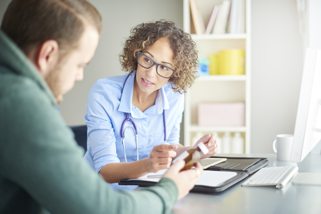 A female doctor wearing glasses and a stethoscope is sitting at a desk, attentively speaking with a male patient.