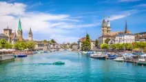 Scenic Swiss city riverfront with churches, bridges, boats, and a bright blue sky.