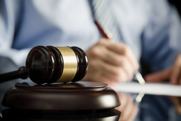 A close-up image of a wooden judge's gavel with a golden band resting on a sound block on a reflective surface.
