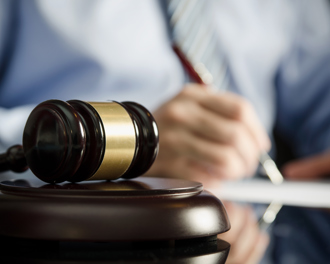 A close-up image of a wooden judge's gavel with a golden band resting on a sound block on a reflective surface.