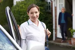  Smiling home carer in uniform arriving by personal vehicle, standing by the car outside a client’s house