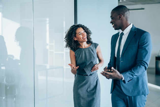 Two professionally dressed colleagues walking and chatting in a bright modern office corridor, smiling and engaged in conversation
