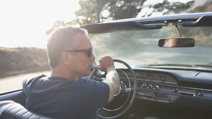 Man wearing sunglasses driving a classic convertible on a sunny day, viewed from the passenger seat